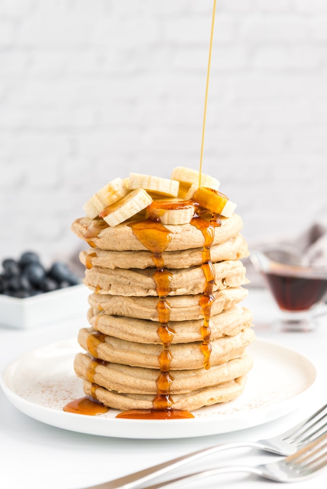 fluffy vegan pancakes stacked and topped with sliced bananas and blueberries, and maple syrup pouring over top